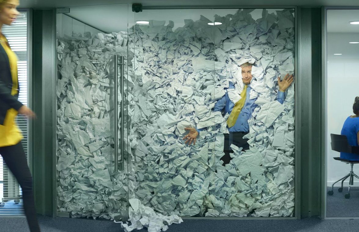 A businessman is trapped in his glass office by a surplus of discarded ideas on paper . His colleague in the next office is working more efficiently and is oblivious to him being trapped , as is a passing female office worker